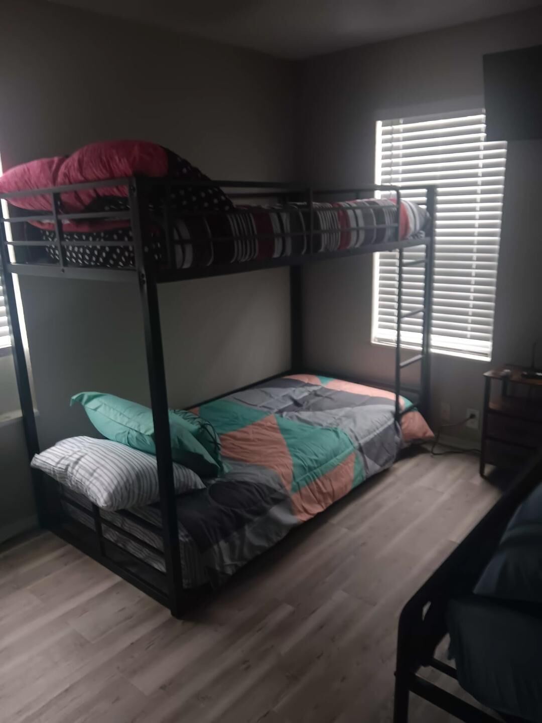 A bunk bed with a patterned blanket and pillows, a desk with a chair, and a window with blinds.