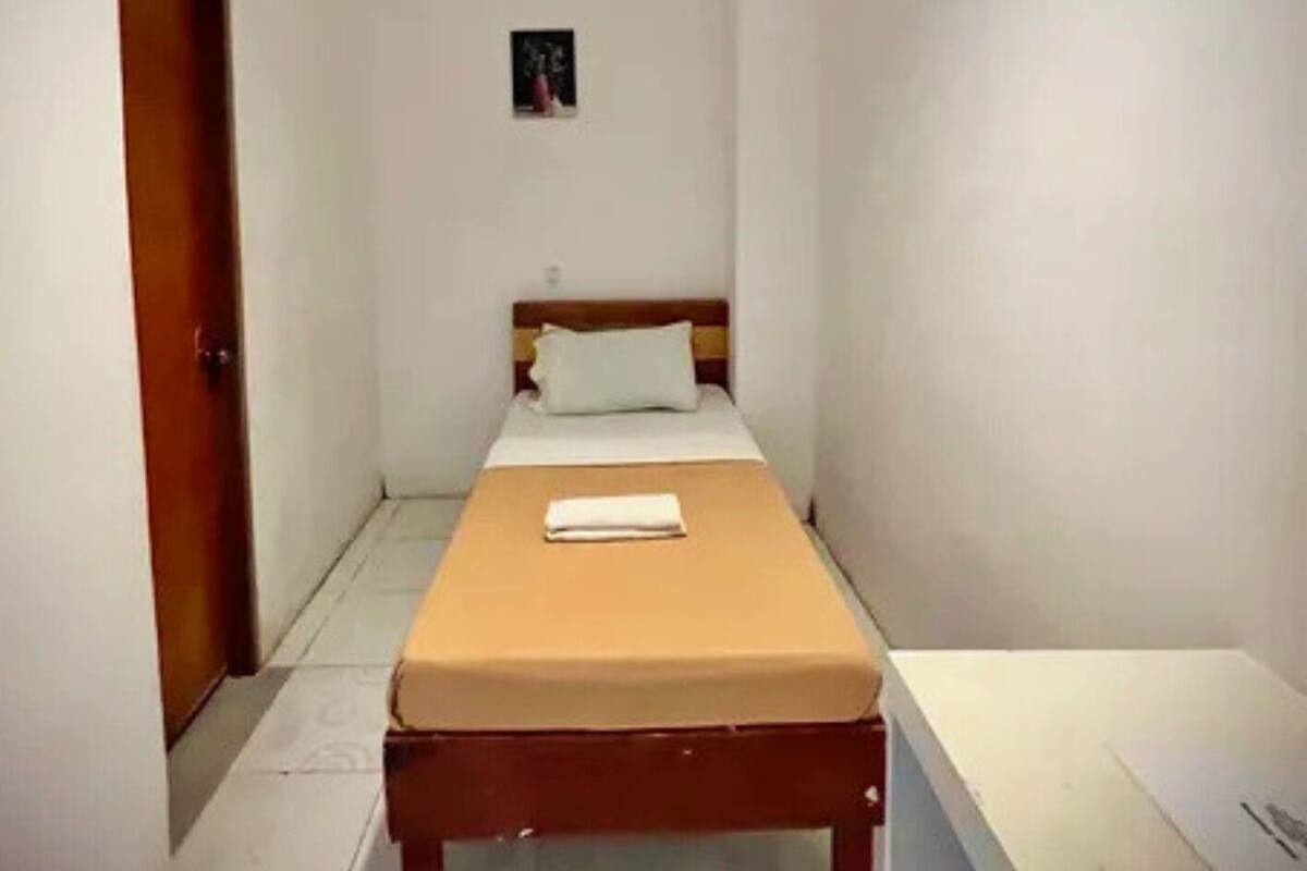 A small, narrow room with a single bed, a desk, and a door.
