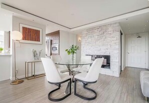 A modern dining area with a glass table and white chairs, a stone fireplace, and a minimalist kitchen in the background.