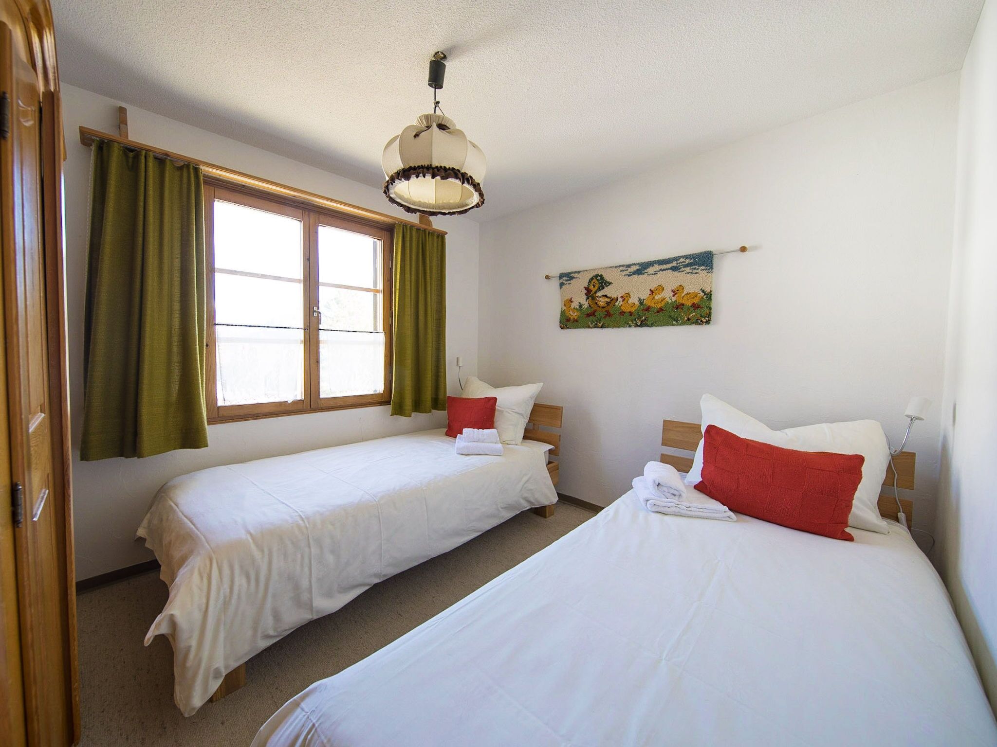 A room with two single beds, a window with green curtains, a wall-mounted lamp, and a painting.
