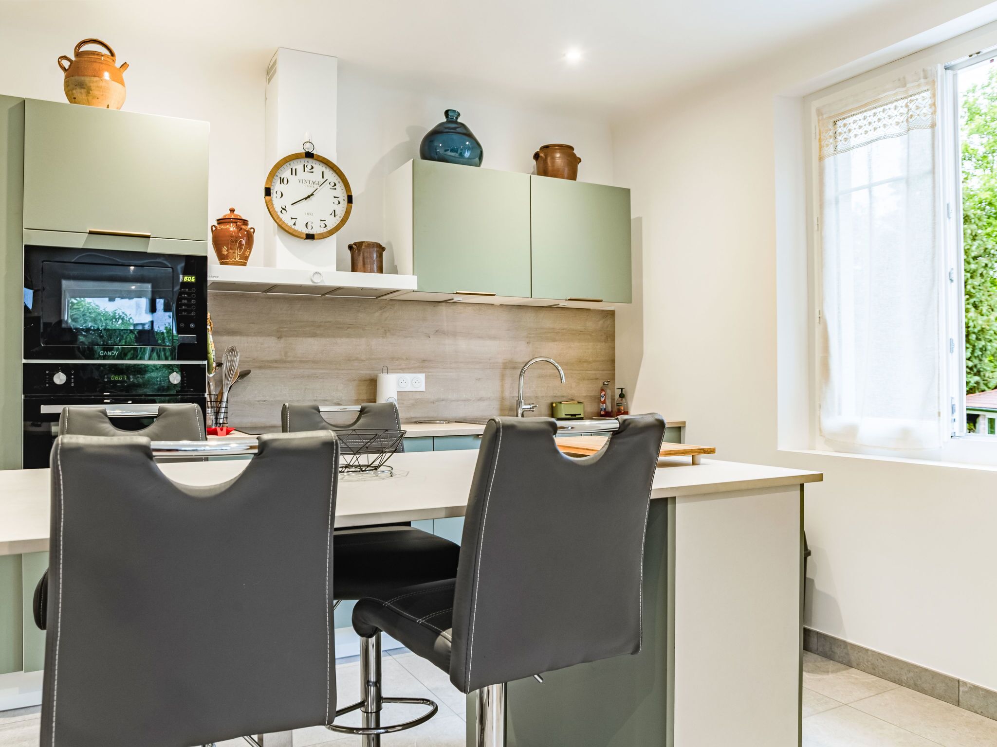A modern kitchen with a built-in oven, a clock, and a window with a view of greenery.
