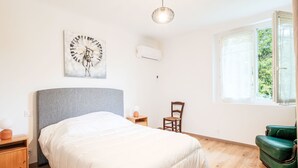 Un dormitorio con cama, una silla, una mesita, un reloj de pared y una ventana con vista a zonas verdes.