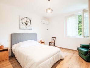 Ein Schlafzimmer mit Bett, Sessel, kleinem Tisch, Wanduhr und Fenster mit Blick auf Grünfläche.