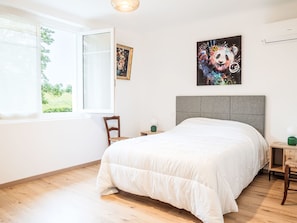 Ein Schlafzimmer mit Bett, Sessel, Nachttisch, Fenster und Wandbildern.