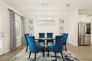 Ein Essbereich mit einem Glastisch, blauen Polsterstühlen, einer Chandelüster und einem Edelstahlkühlschrank.