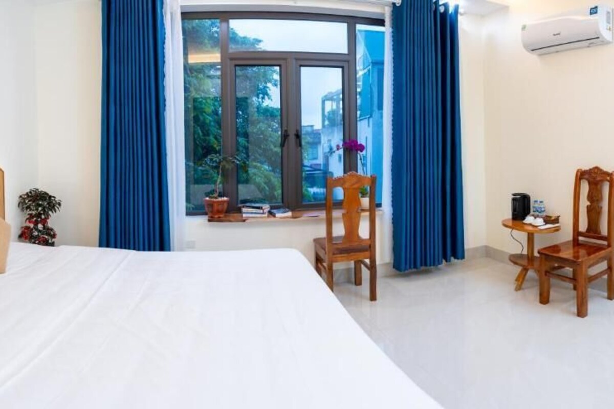 A bedroom with a wooden headboard, a large bed, two chairs, a small table, a potted plant, and a window with curtains.