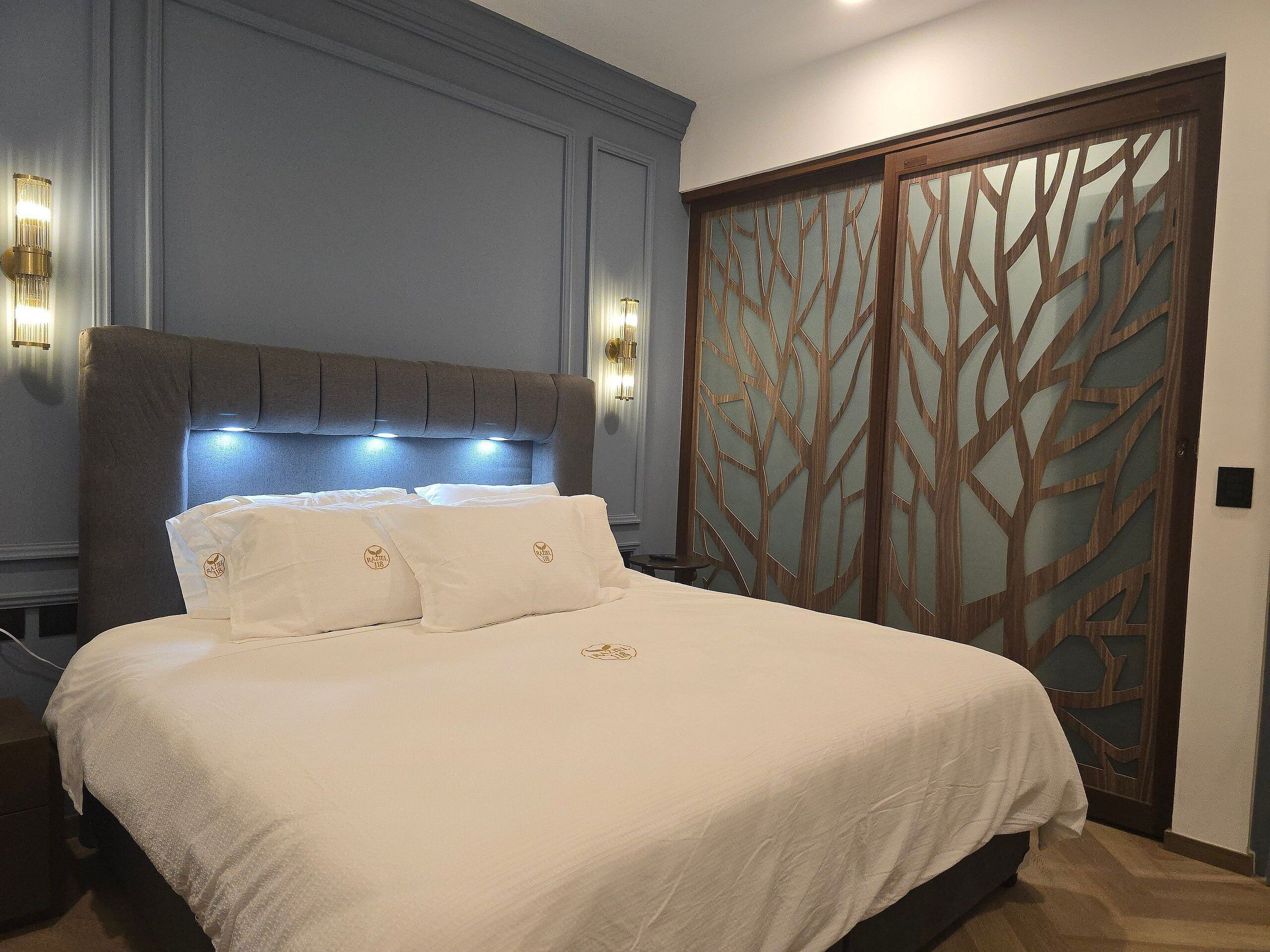 Una cama bien hecha con ropa blanca y almohadas, un cabecero con iluminación integrada y una puerta de madera decorativa con un motivo geométrico.