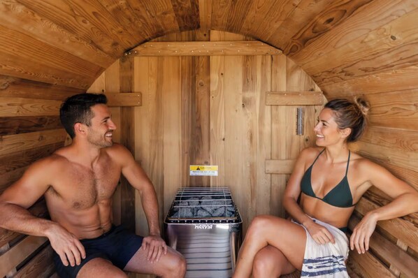 A wooden sauna with two people sitting inside, a Harvia sauna heater, and a caution sign.