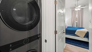 A compact room with a washing machine, a bed with a blue bedspread, a ceiling fan, and a door leading to another room. - Luxury Retreat on Oleander - Unit J (Dauphin Island)