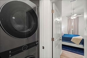 A compact room with a washing machine, a bed with a blue bedspread, a ceiling fan, and a door leading to another room.