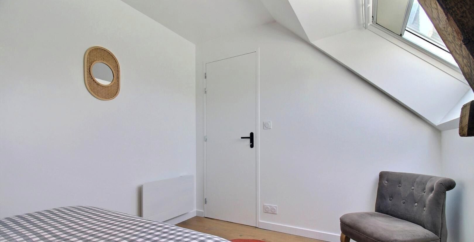 A room with a bed, a chair, a door, a window, and a wall-mounted mirror.