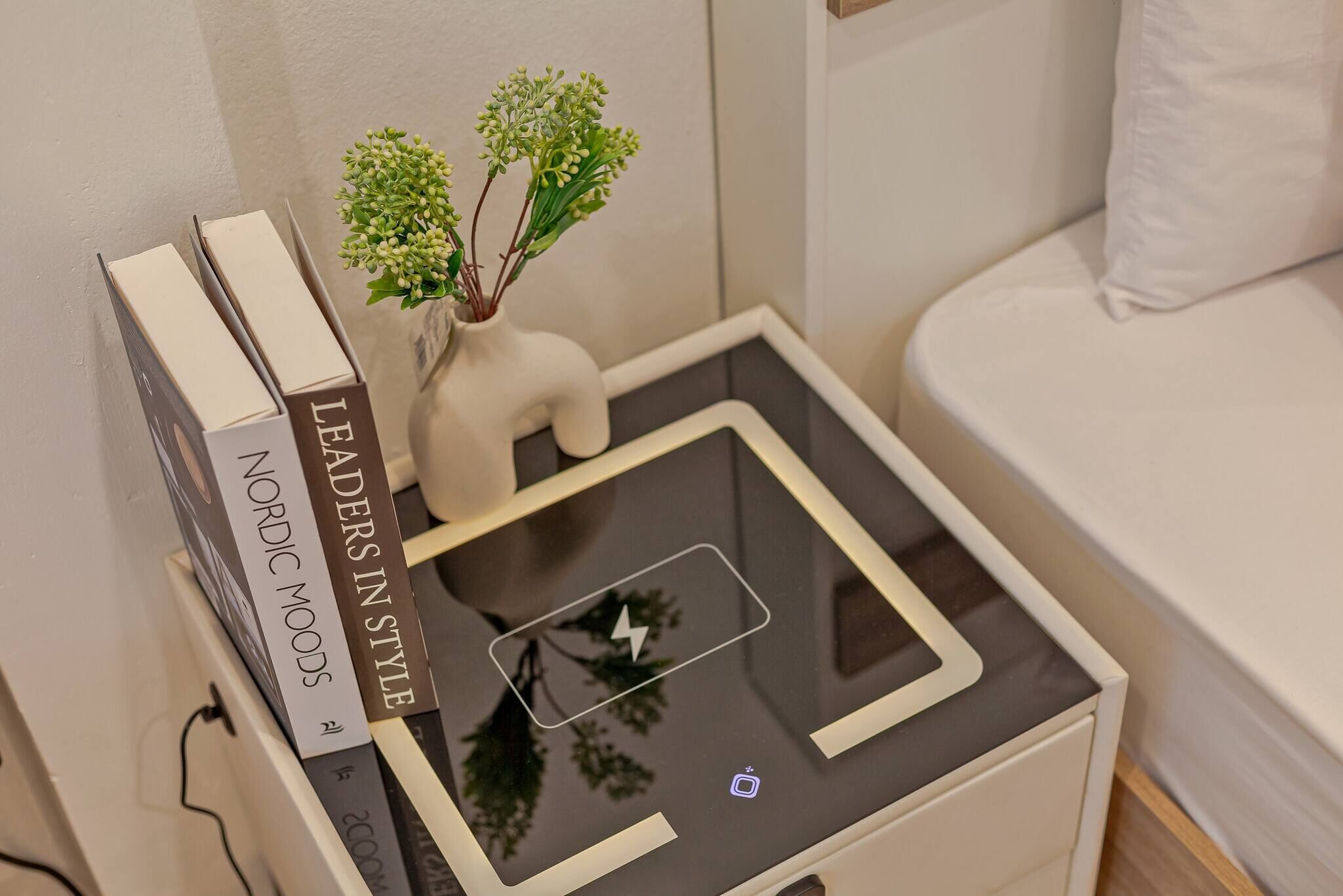 A bedside table with a book titled 'Leaders in Style,' a small vase with green plants, and a smartphone on the table.