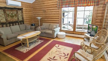 A log cabin interior with a sofa, a small table, a wicker chair, a television, and a large window.