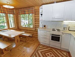 Puinen mökki, jonka sisätiloissa on keittiö, ruokapöytä, penkit ja suuret ikkunat.