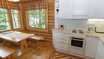 A wooden cabin interior with a kitchen, dining table, benches, and large windows.