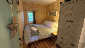 Ein Zimmer mit Holzwänden, einem Bett, einem Nachttisch, einem Kleiderschrank und einem Fenster mit Vorhängen.