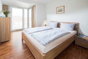 Une chambre moderne avec un lit en bois, une table de chevet, une armoire et une grande fenêtre donnant sur des immeubles.