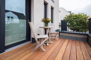 Eine Holzterrasse mit zwei Stühlen und einem Tisch, einer Vase mit Blumen und Blick auf ein Gebäude durch ein Fenster.