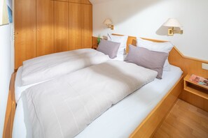 Ein Doppelbett mit weißer Bettwäsche, ein Holzschrank, zwei Wandlampen und ein kleiner Nachttisch mit einer Zeitschrift.