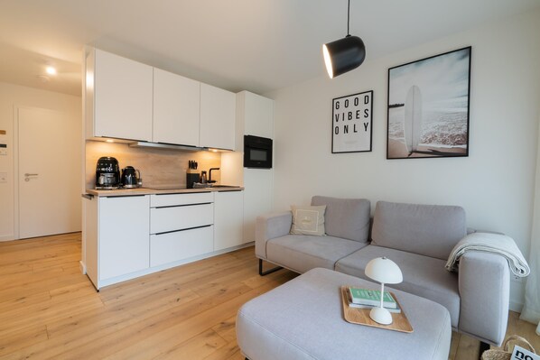 Eine moderne Küche mit weißen Schränken, einer Kaffeemaschine und einer Mikrowelle. Ein graues Sofa und ein kleiner Tisch mit Lampe und Büchern stehen dort. Zwei gerahmte Bilder hängen an der Wand.