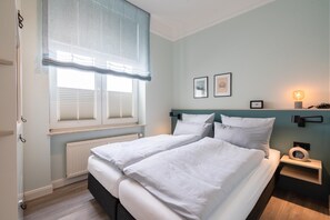 Ein Schlafzimmer mit zwei Betten, einem Fenster mit Jalousien, einem Nachttisch mit Lampe sowie gerahmten Bildern an der Wand.