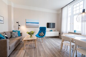 Ein modernes Wohnzimmer mit einer Couch, einem blauen Sessel, einem Esstisch, einem Fernseher und einem Fenster mit Vorhängen.