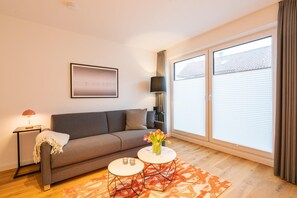 Ein modernes Wohnzimmer mit einer Couch, zwei Beistelltischen, einer Lampe, einer Blumenvase und großen Fenstern mit Jalousien.