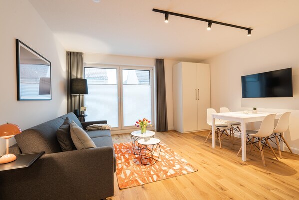 Ein modernes Wohnzimmer mit einer Couch, Couchtisch, Esstisch und Stühlen. Ein Flachbildfernseher ist an der Wand befestigt, auf dem Beistelltisch steht eine Lampe und es gibt ein Fenster mit Vorhängen.