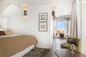 Quarto de hotel com uma cama, uma cadeira, uma mesa com um copo e duas imagens emolduradas na parede.