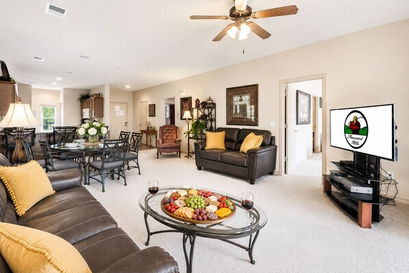 A spacious living room with a leather sofa, a glass coffee table with fruit and wine, a dining area with chairs, a leather couch, and a television. - Branson Condo with Golf Course View • 3 King Suites • Sleeps 6 (Branson)