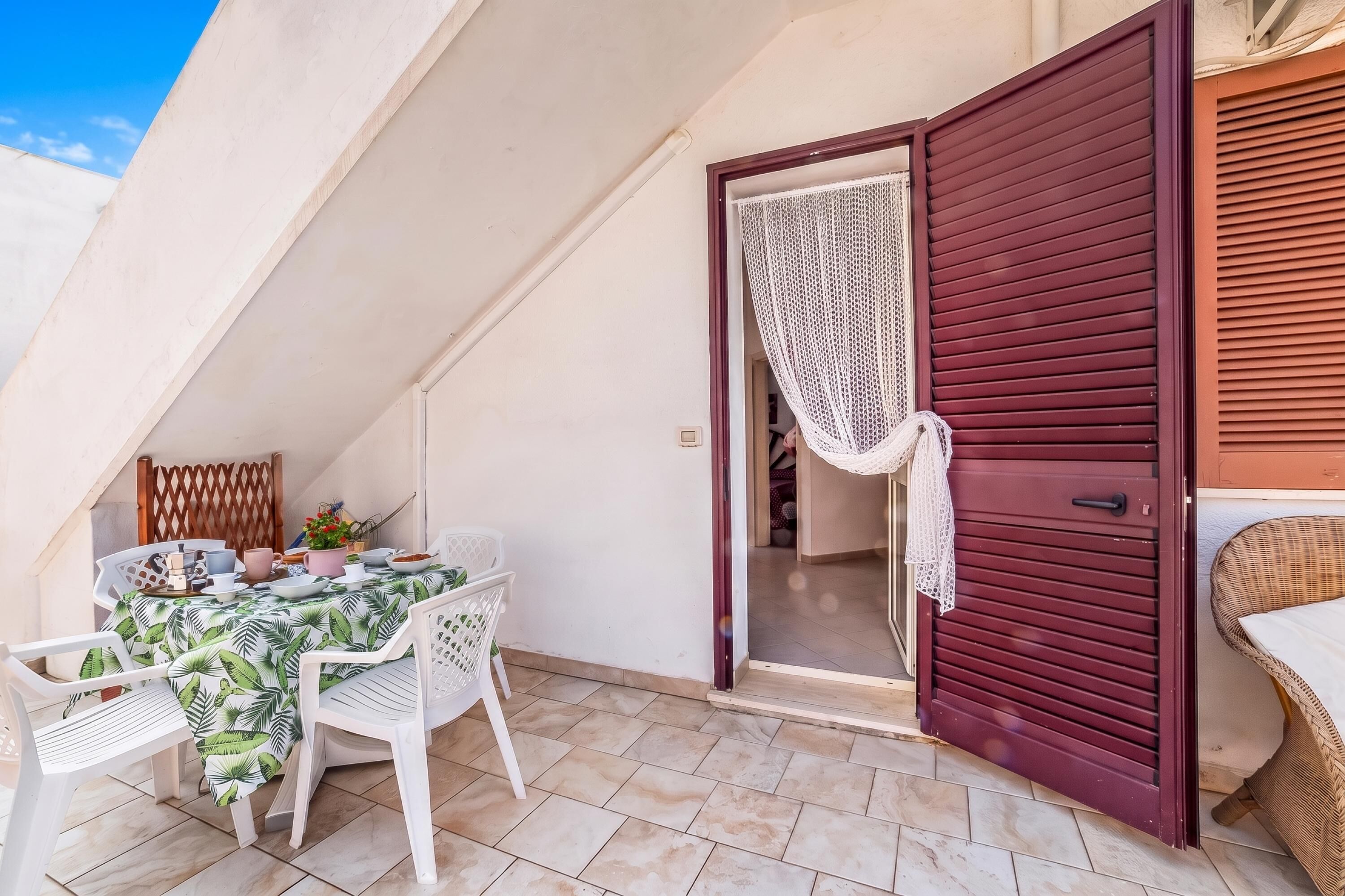 A balcony with a table set for dining, white plastic chairs, and a red door with a white curtain.