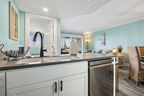 A modern kitchen with a sink, dishwasher, and a dining area with a wicker chair and a lamp. - Partial Ocean-view condo with 2 bedrooms, perfect for a beach getaway (Fernandina Beach)