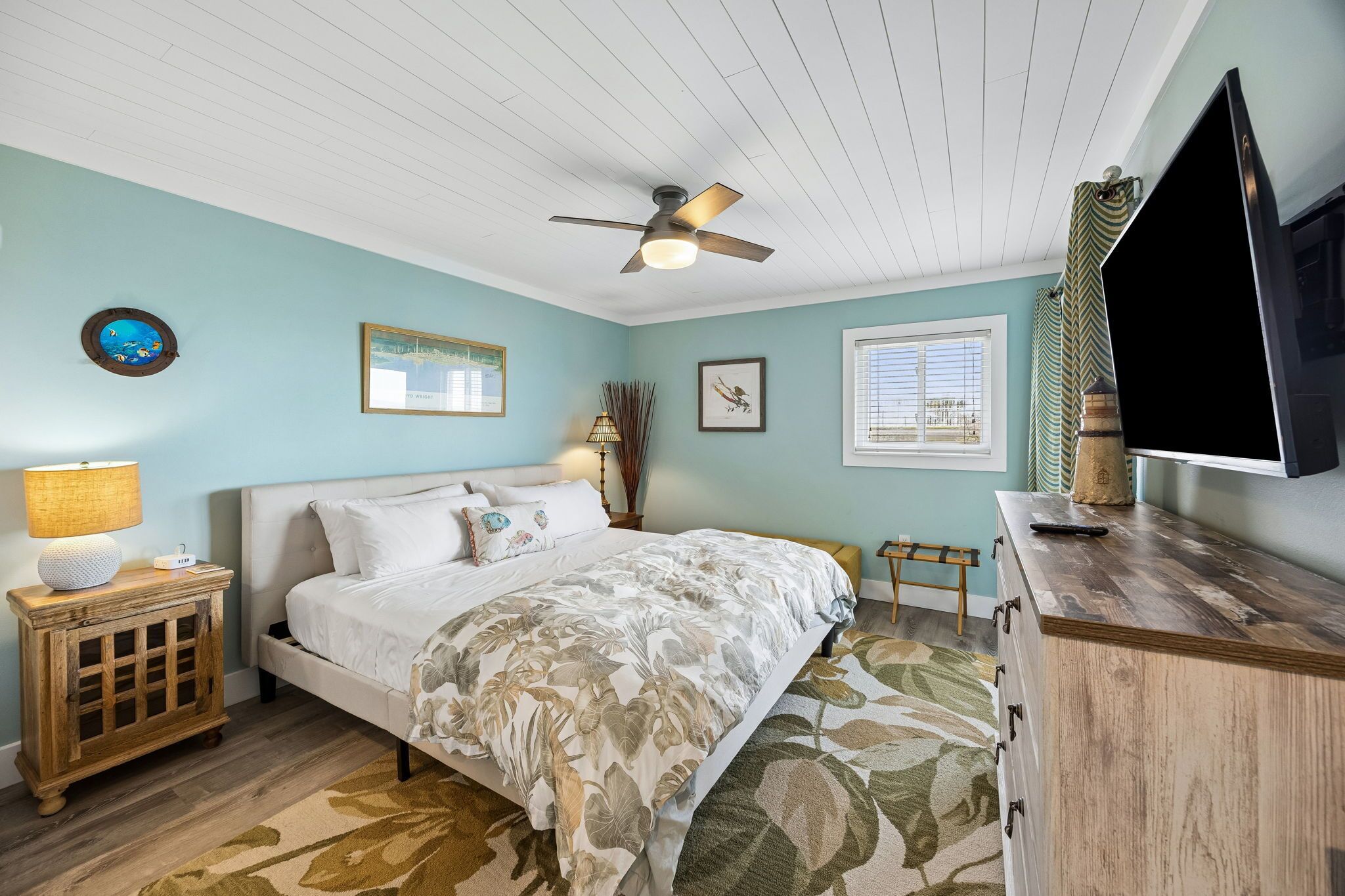 A bedroom with a bed, a ceiling fan, a TV, a nightstand, and a window with blinds.