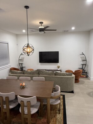 Ein modernes Wohnzimmer mit einem Ecksofa, einem Esstisch mit Stühlen, einem Deckenventilator, einem Wand-Fernseher und einer dekorativen Leuchte.