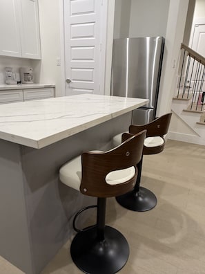 Eine moderne Küche mit weißer Marmorarbeitsplatte, Edelstahlkühlschrank und zwei Barhocker mit Holzsitzen und schwarzen Füßen.