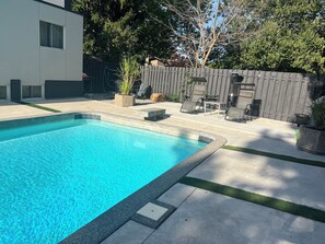 A rectangular swimming pool with a tiled edge, surrounded by a concrete patio with a small grassy area.