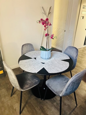 Uma mesa redonda com toalha branca texturizada, quatro cadeiras estofadas cinzas e um vaso com flores cor-de-rosa.