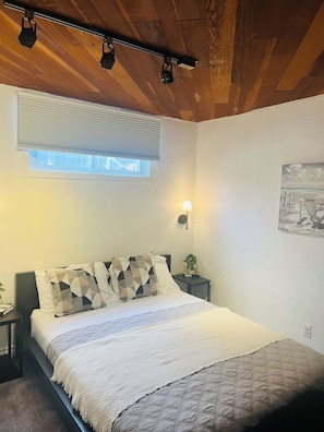 Une chambre à coucher avec un lit, une fenêtre avec des stores, une lampe fixée au mur et un tableau.
