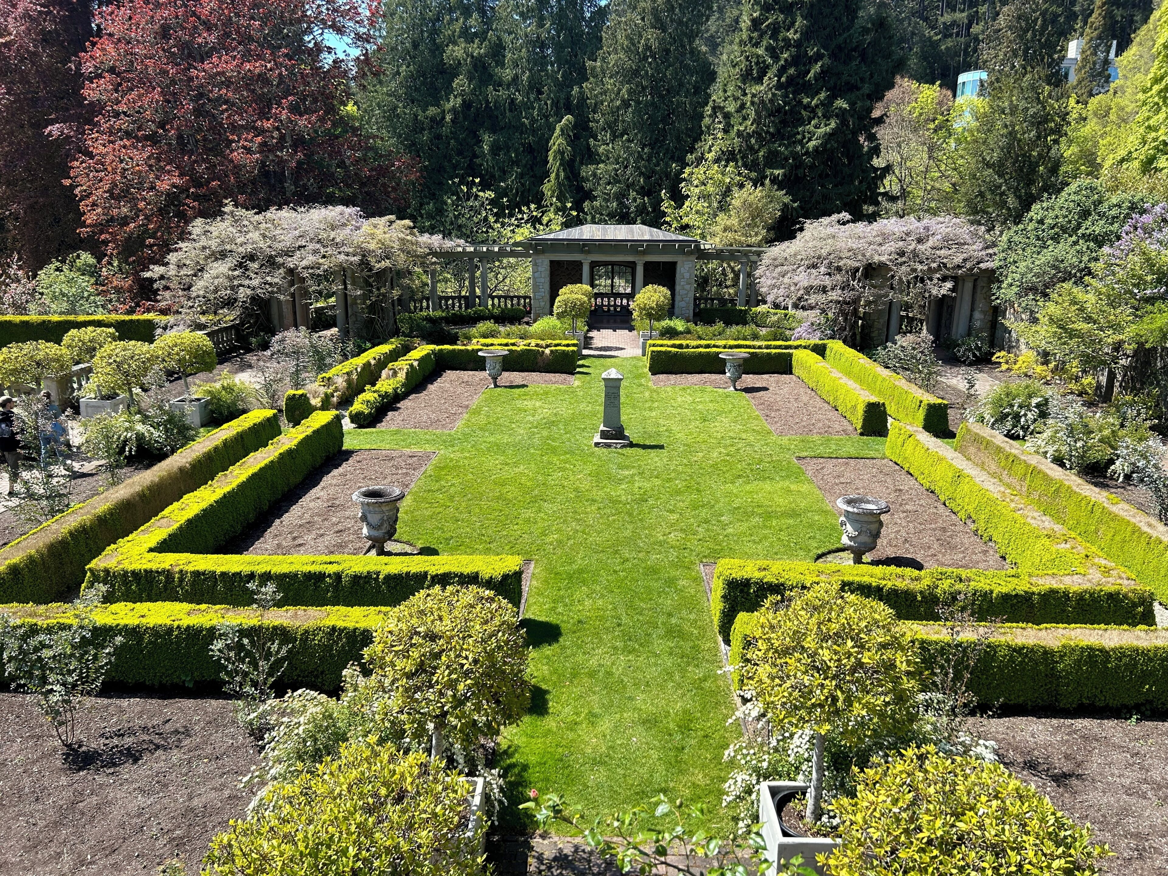 A formal garden with geometrically trimmed hedges, a central pavilion, and stone urns.
