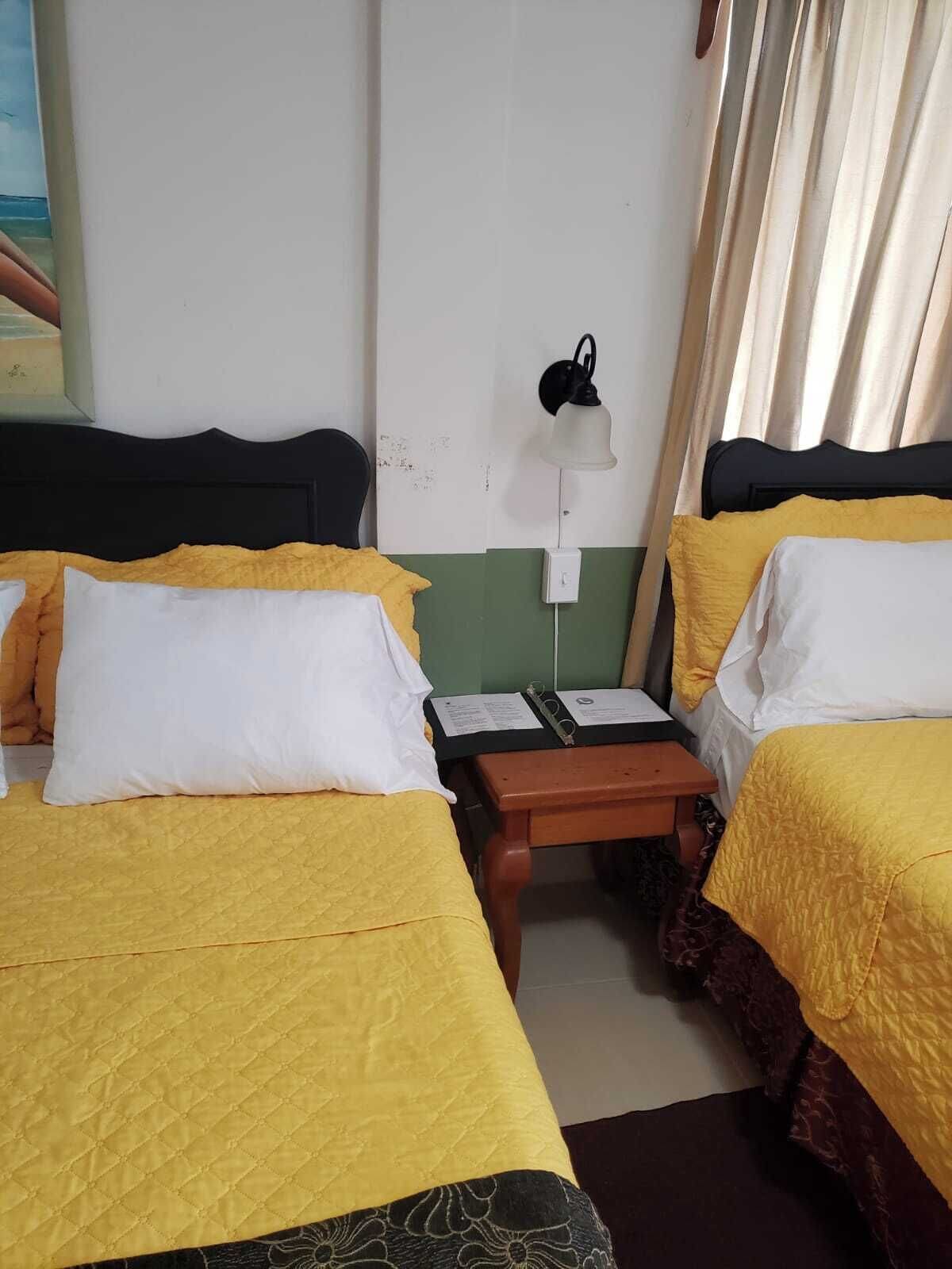 A room with two beds, each with a yellow bedspread and white pillows, a small wooden nightstand with a lamp, and a wall-mounted light.
