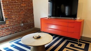 A room with a brick wall, a red dresser with drawers, a flat-screen TV on top, and a round table with a plate and cup. - Brick & Bed, Modern Loft, Walk to Arch, Stadium, 402 (St. Louis)