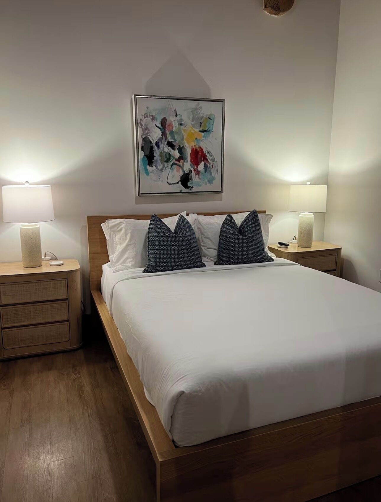 A neatly made bed with white linens, two patterned pillows, a wooden headboard, and two bedside tables with lamps.