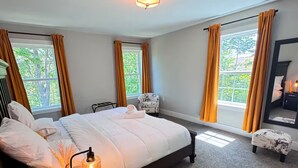 Ein Schlafzimmer mit einem großen Bett, zwei mit Vorhängen versehenen Fenstern, einem Sessel, einem Nachttisch mit Lampe und einem Spiegel an der Wand.