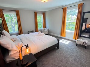 Ein Schlafzimmer mit einem großen Bett, zwei mit Vorhängen versehenen Fenstern, einem Sessel, einem Nachttisch mit Lampe und einem Spiegel an der Wand.
