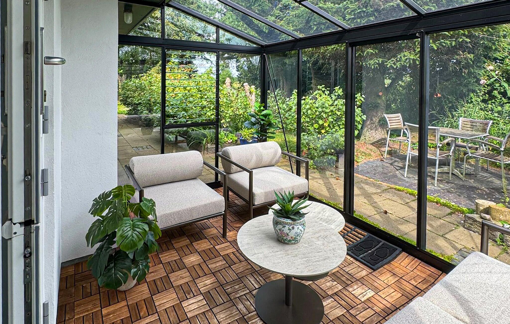 A conservatory with a wooden floor, two cushioned chairs, a small table, and a potted plant.