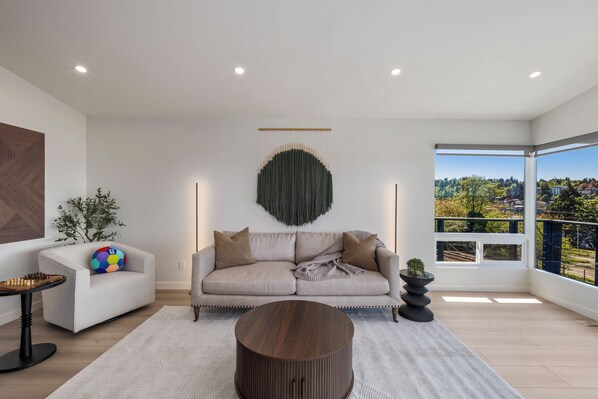 A modern living room with a sofa, a round coffee table, and a view of a cityscape through large windows.