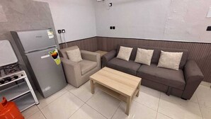 Een woonkamer met een bank, een bijzettafeltje, een koelkast en een keukenhoek.