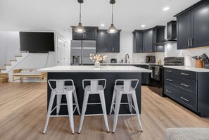 A modern kitchen with dark cabinets, a large island, and three bar stools. - 4BR Spacious Home with Deck, BBQ,Fire Pit,Sleeps 8 (Arvada)