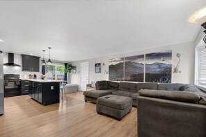 A modern kitchen with dark cabinetry, a central island, pendant lighting, and a large window. - 4BR Spacious Home with Deck, BBQ,Fire Pit,Sleeps 8 (Arvada)