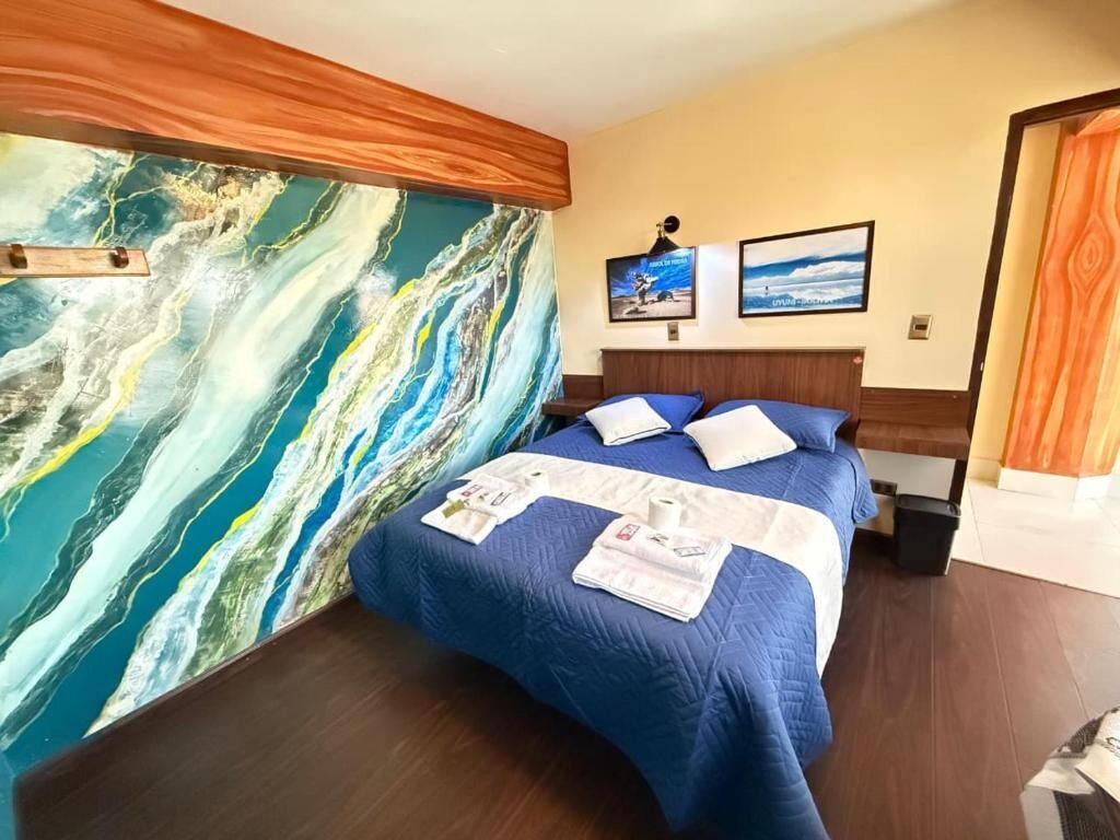A hotel room with a large abstract painting on the wall, a bed with blue bedding, and two framed pictures on the wall.